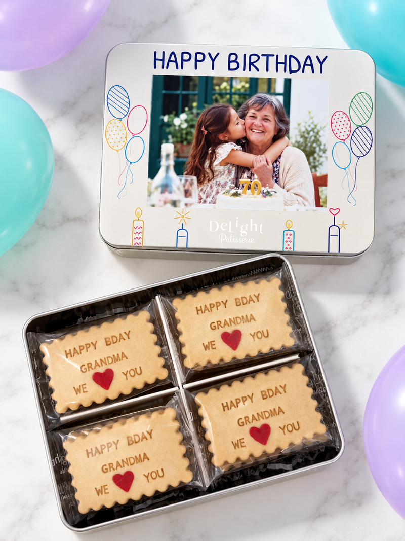 A personalized birthday cookie tin gift with a printed photo lid that says 'HAPPY BIRTHDAY' and a separate tin holding four custom shortbread cookies printed with 'HAPPY BDAY GRANDMA WE ❤️ YOU'. 