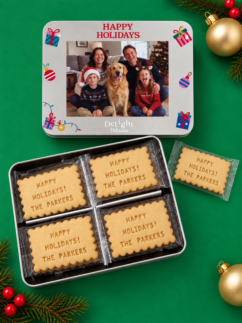 A happy holiday cookie tin set on a green background with Christmas ornaments and evergreen branches. The set includes a silver tin with a printed family photo and 'Happy Holidays' message, an open tin showing four custom message shortbread cookies.